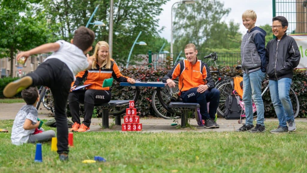 Speeltuintje in de buurt. twee buurtsportcoaches met sportkleding aan en 4 kinderen. 1 kind is blikken aan het omgooien met een pittenzak. De kinderen kijken blij.