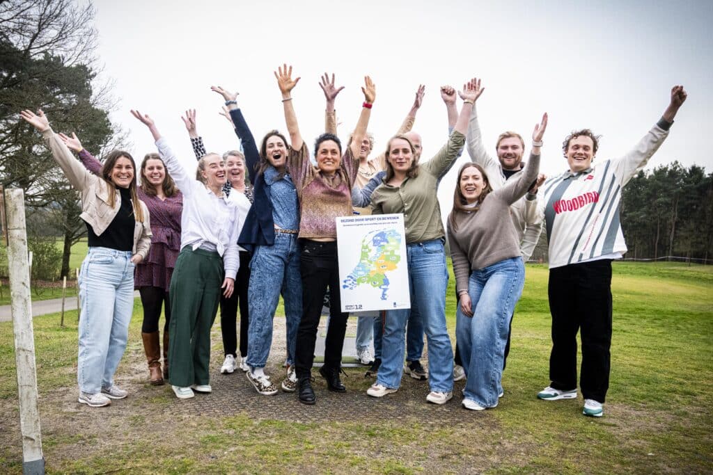 meer dan 10 personen die met hun handen in de lucht op de foto staan. ze hebben een bord vast met de kaart van nederland en de tekst: gezond door sport en bewegen. Domeinoverstijgende samenwerking voor gezonde regio's.