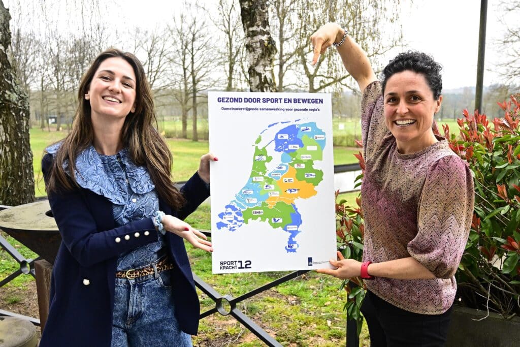 Twee lachende vrouwen (links Sanne, rechts Rosa) met een bord in hun handen. op dit bord staat de kaart van nederland en de tekst 'gezond door sport en bewegen. Domeinoverstijgende samenwerking voor gezonde regio's'