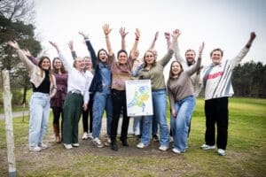 meer dan 10 personen die met hun handen in de lucht op de foto staan. ze hebben een bord vast met de kaart van nederland en de tekst: gezond door sport en bewegen. Domeinoverstijgende samenwerking voor gezonde regio's.