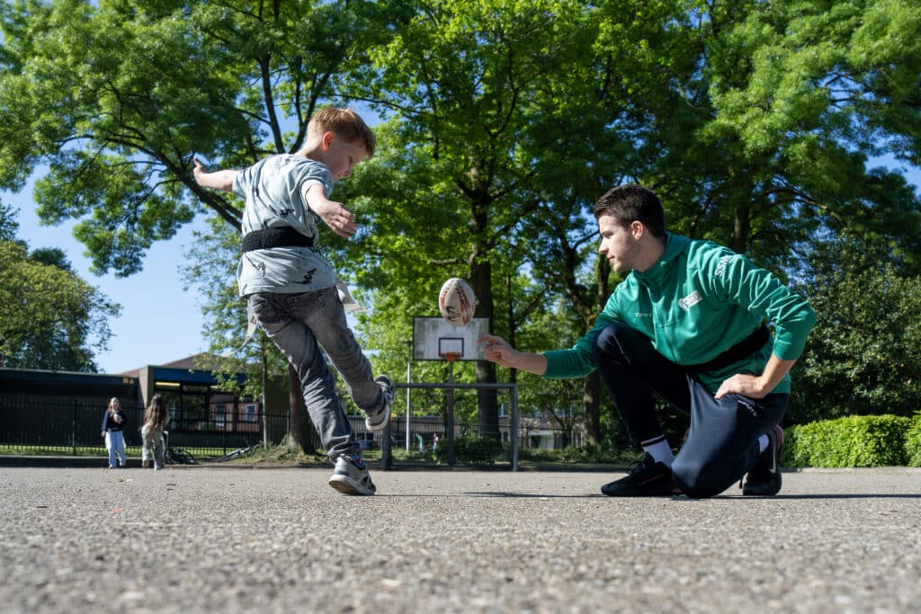 buurtsportcoach ondersteunt jongere bij het trappen van een rugbybal