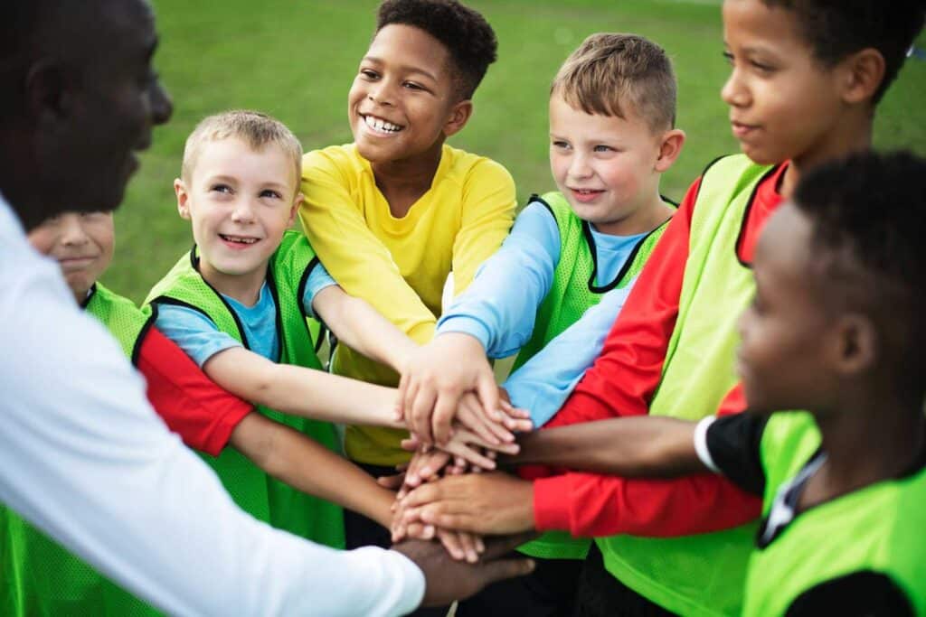 5 kinderen en een volwassenen die hun handen op elkaar hebben en lachen. Ze staan op een sportveld