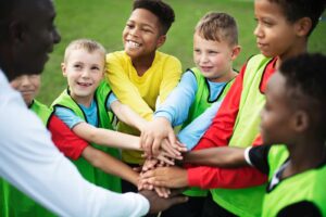 stockfoto van 5 kinderen en een volwassenen die hun handen op elkaar hebben en lachen. Ze staan op een sportveld