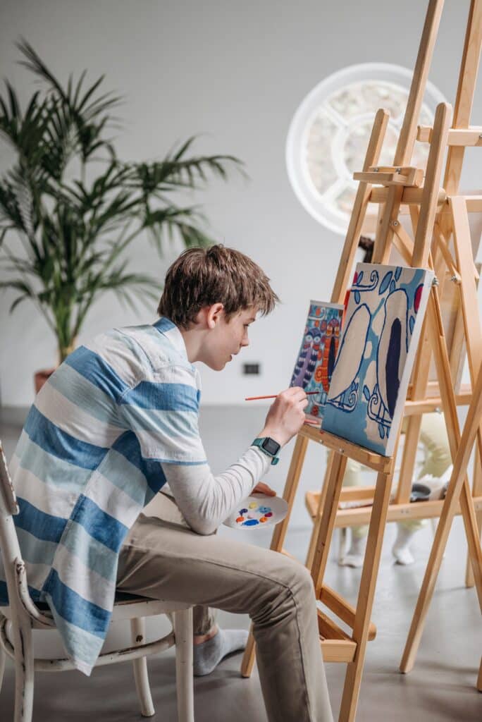 jongen zit op kruk voor schildersezel. Hij is een schilderij aan het schilderen