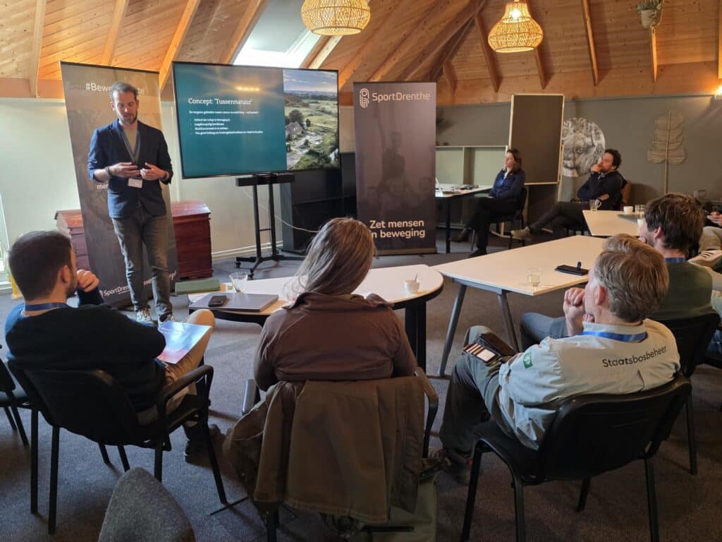 volwassenen zitten op stoelen in een kring. Ze kijken allemaal naar een scherm waarop een presentatie te zien is. Er staat een man voor de groep te vertellen.